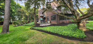 Back yard of large Tudor style brick home with natural landscaping design. Black mulch bed wrapping around home with beautiful green perennial plants, shrubs and trees.