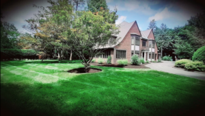 Amazing Tudor style brick home with lush green mowed grass and landscaping surrounding the house. Yard bordered by tall hedge row with trees.
