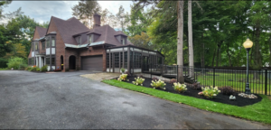Front of large stunning Tudor style home displaying landscaping with flowering shrubs planted in black mulch bed. Tall trees and black gate background.
