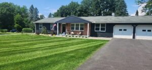 Blue ranch house with striped lawn leading to landscape bed on sunny day. Green, purple and yellow shrubs in front of home.